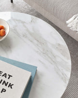 A round marble-top coffee table with a sleek modern design, accompanied by a light grey fabric sofa with a plush texture and a fringed throw pillow. A grey textured rug underpins the setting, and a book with a turquoise cover adds a pop of colour to the scene.