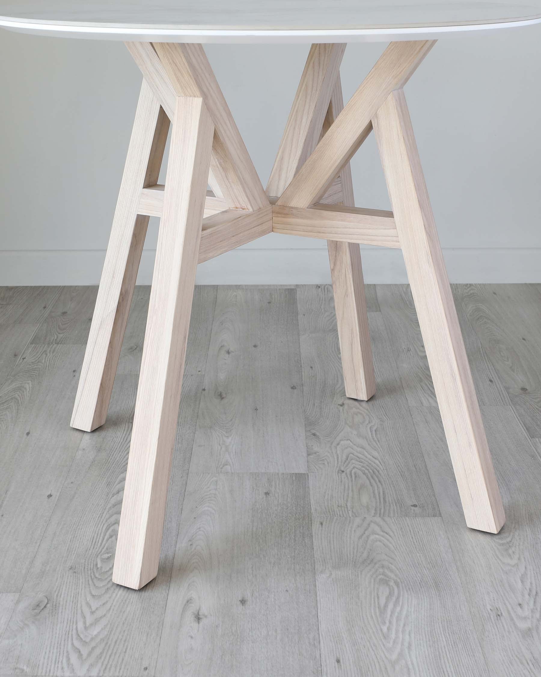 Modern light wood table with a round white top and unique crisscross leg design, set on a grey wood flooring.