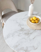 A modern round white marble-top dining table with a smooth finish and a grey veining pattern, paired with a single sleek taupe dining chair with a curved backrest. The table is accessorized with a circular braided placemat, a white textured vase holding pampas grass, and a shallow bowl with fresh lemons. The chair appears to have wooden legs that match the light hardwood flooring, creating a harmonious and minimalist aesthetic.