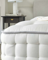 Close-up of a plush white tufted mattress with detailed stitching and a grey piping edge, next to a dark wood nightstand with a brass knob, featuring a white lamp with a cylindrical shade, a stack of books, and a clear drinking glass.