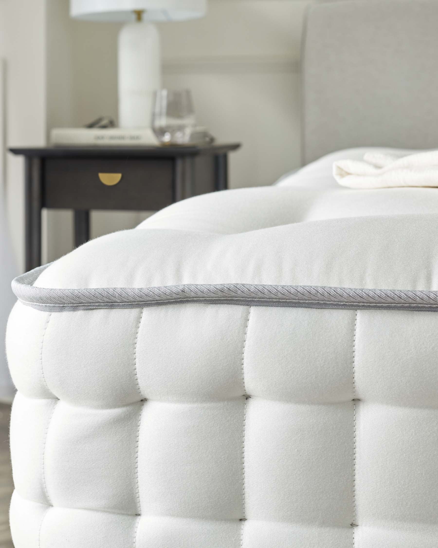 Close-up of a plush, tufted mattress with a grey piping detail, paired with a sleek, dark wooden bedside table featuring a single drawer with a brass handle, and a contemporary white ceramic lamp with a white shade on top.