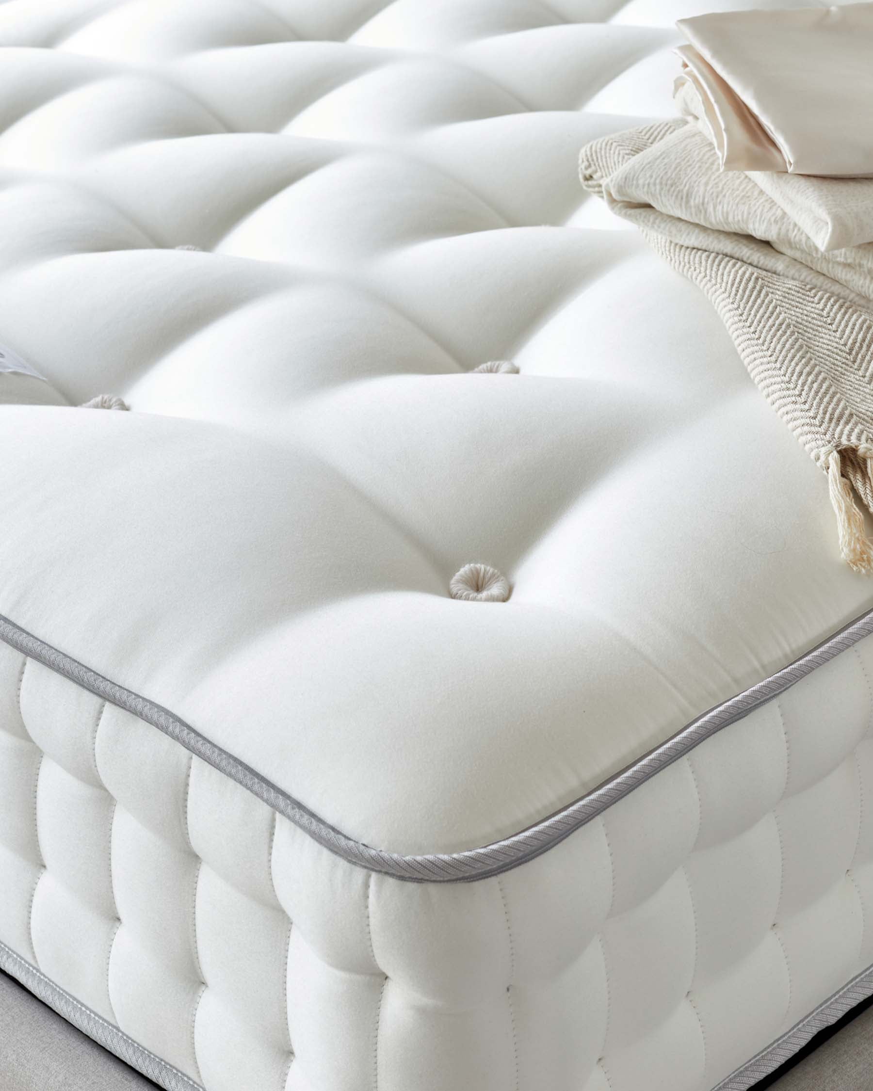 Close-up of an elegant, cream-colored tufted ottoman with a plush, buttoned top cushion and a knitted throw and accent pillow resting on it, featuring a detailed grey piping along the edge.