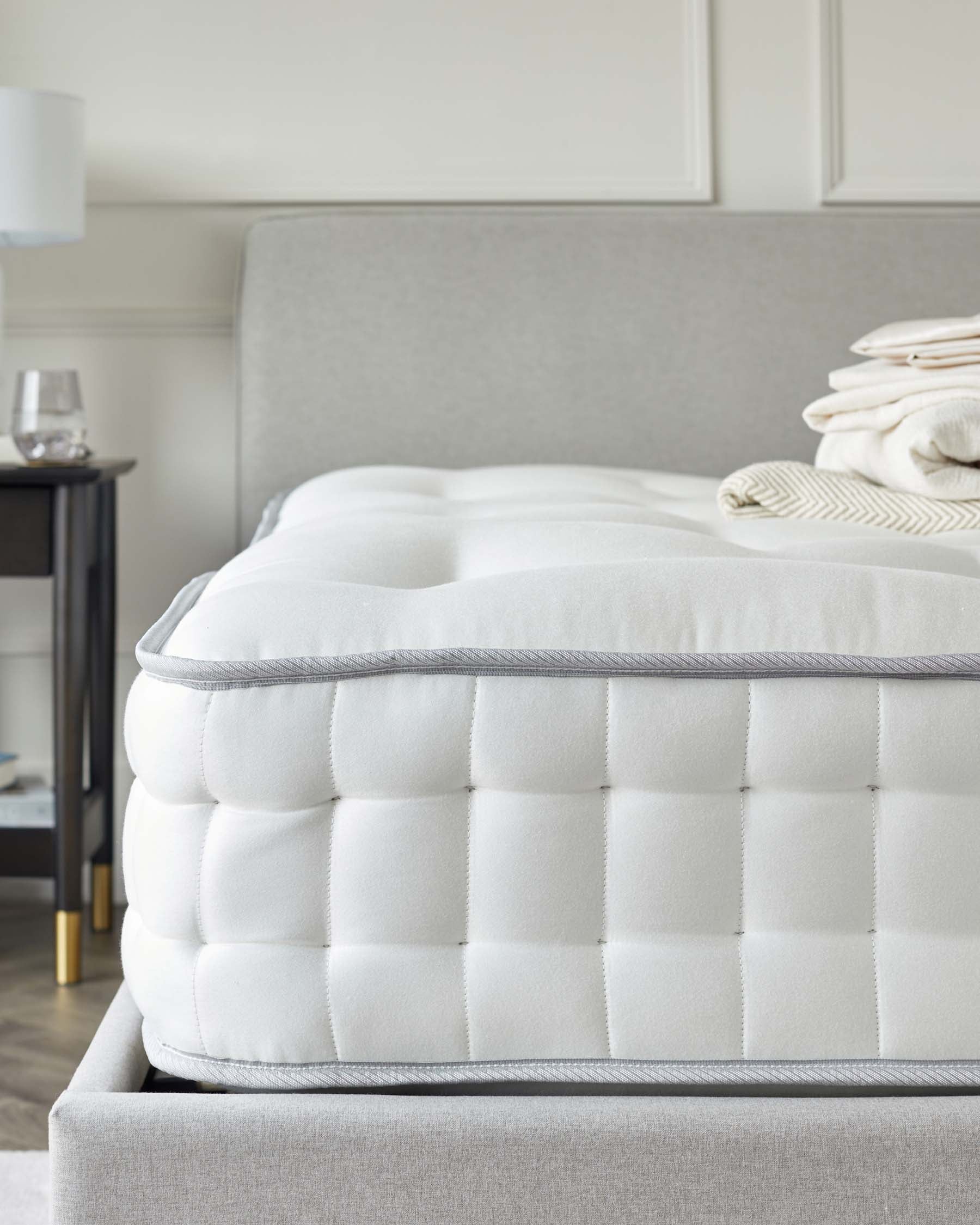 Close-up of a plush, tufted mattress with white bedding atop a grey upholstered bed frame, paired with a dark bedside table featuring a white lamp and clear glass. The room has a neutral colour palette, conveying a sense of luxury and comfort.