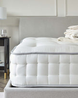 Close-up view of a luxurious, tufted mattress on a bed frame with a light grey upholstered headboard. The bed is accessorized with neatly folded, cream-colored linens and subtly patterned throw blanket. In the background, a small round bedside table with a dark finish and thin metal legs, paired with a simple, modern table lamp with a white shade, can be seen. The room has a muted, sophisticated colour palette.