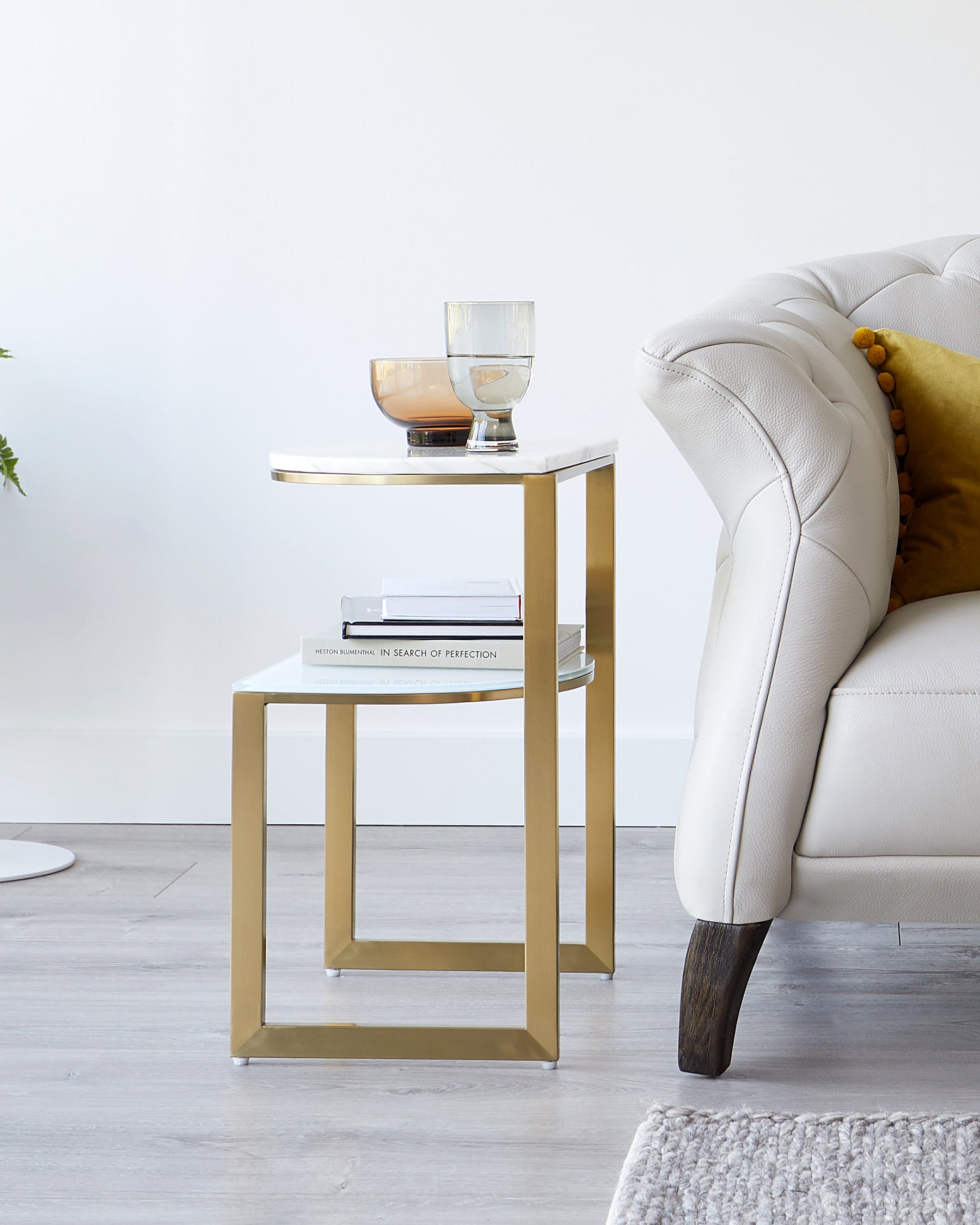 A modern gold metal side table with a square top and a base that doubles as a lower shelf, adorned with decorative glassware and books, next to a cream tufted armchair with a dark wooden leg visible and a mustard yellow throw pillow.