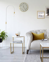 Elegant living room setup featuring a white tufted armchair with dark wooden legs and golden button accents, paired with a mustard yellow throw pillow. Beside it, a modern two-tiered round side table with a gold frame and white tabletops holds books and a glass bowl, complementing a white lamp with a curved stand and gold accents. A grey area rug lies partially underneath, adding texture to the light wooden flooring.