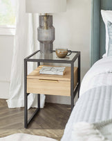 Modern bedside table with a black metal frame and natural wood finish drawer, accessorized with a decorative lamp and a coffee cup on its surface, set against a light bedroom interior.