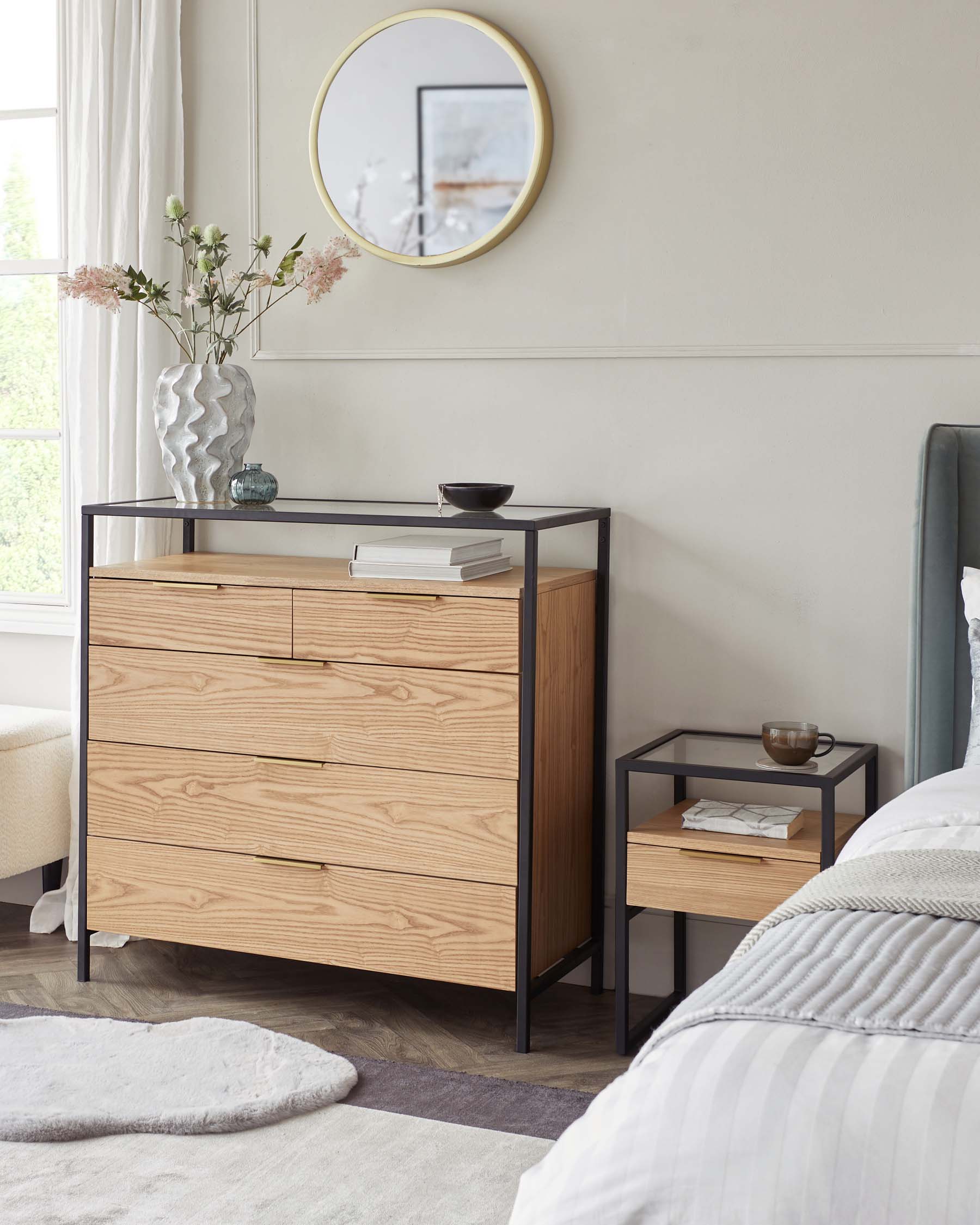 Modern bedroom furniture including a light wood dresser with three drawers and gold handles, and a small matching bedside table with a lower shelf and a single pull-out drawer. Both pieces feature a minimalist design with sleek black metal frames. A circular gold-trimmed mirror hangs on the wall above the dresser.