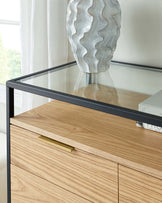 Modern light wood veneer sideboard with a minimalist brass handle, adjacent to a sleek black-framed glass-top side table, in a bright room with a white curtain background.