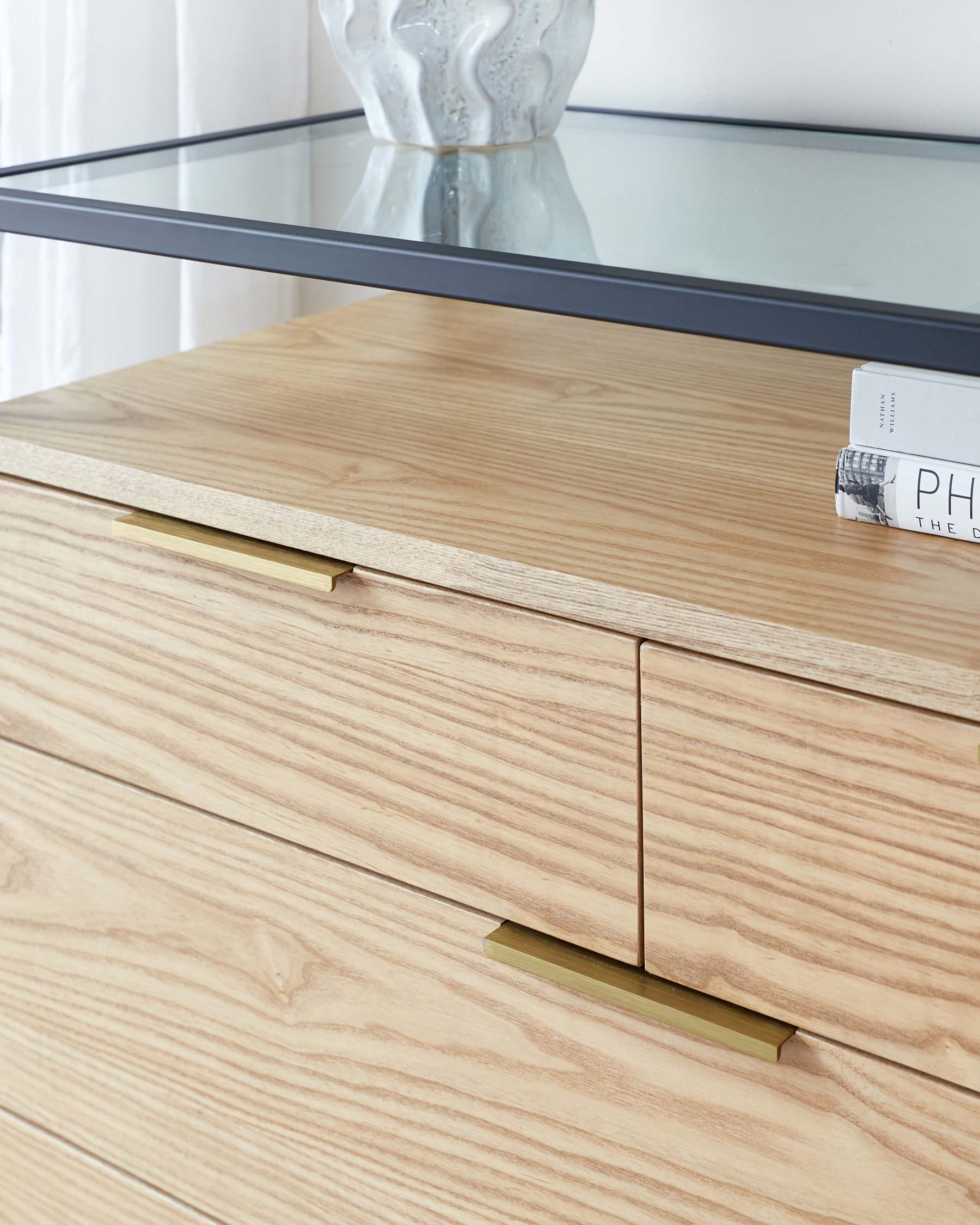 Modern light wood dresser with clean lines and minimalist handles under a glass console table displaying a ceramic vase.