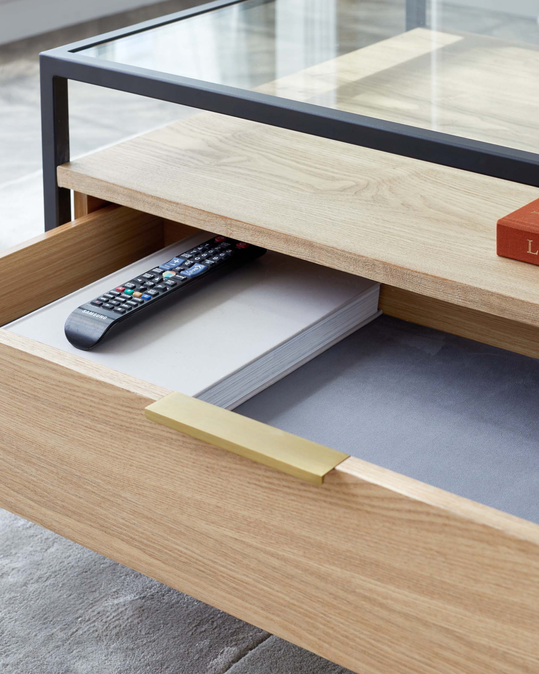 Modern light oak coffee table with a transparent glass top and a sleek black metal frame, featuring an open shelf and a drawer with a brushed brass handle pull.