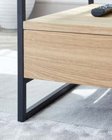 Modern minimalist-style wooden coffee table with a light oak finish and sleek black metal frame, featuring one drawer. The table is placed on a soft grey area rug and holds a remote control.