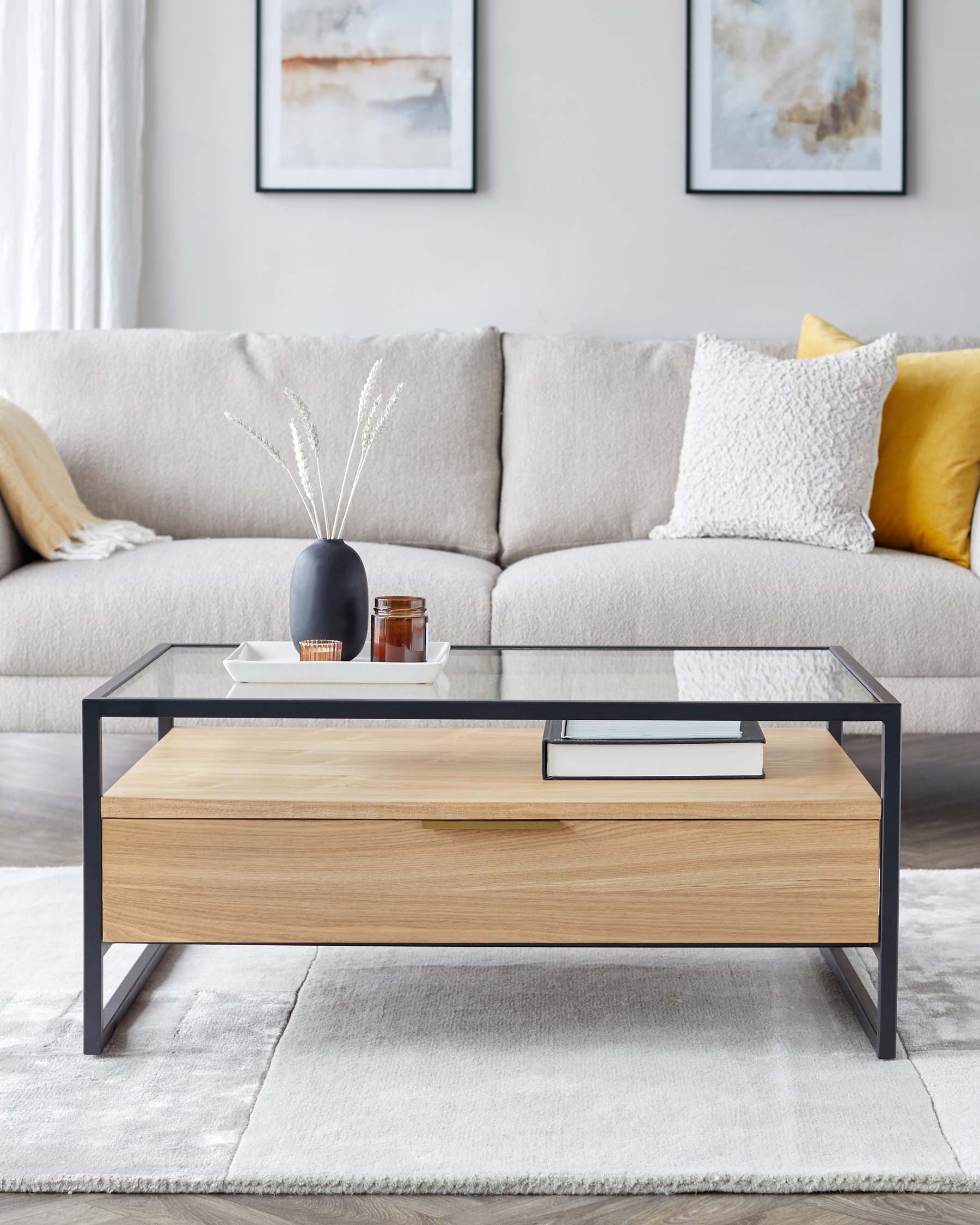 Modern rectangular coffee table with a clear glass top and a black metal frame, featuring a lower shelf with a natural wood finish. The table is accessorized with a decorative vase, books, and a small tray. The background includes a neutral-toned sofa with throw pillows and a framed abstract art piece. The setting is completed with a soft grey area rug.