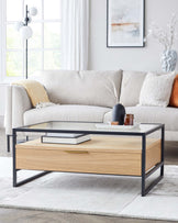 Contemporary rectangular coffee table featuring a black metal frame with a clear glass top and a lower wooden shelf with a natural oak finish. The table is styled with books and decorative items on both tiers.