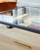 A modern minimalist glass-top coffee table with a wooden base, featuring a simple, elegant design with black metal frames supporting a clear glass surface that reveals the structure underneath.