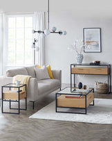 Contemporary living room furniture setup featuring a light grey fabric sofa accented with yellow and white pillows, a rectangular glass-topped coffee table with a wooden drawer and black metal frame, a matching side table with a slimmer profile, and a modern console table with two wooden drawers and an open shelf, also framed in black metal. All pieces showcase clean lines and a minimalist aesthetic, arranged on a soft grey area rug.