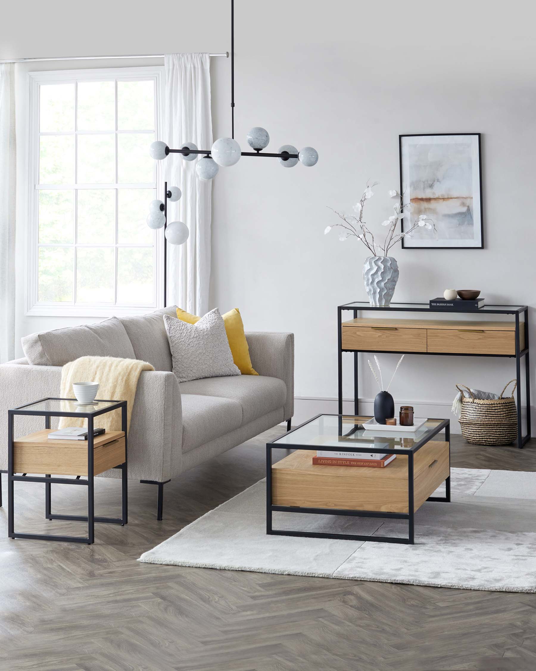 Contemporary living room furniture setup featuring a light grey fabric sofa accented with yellow and white pillows, a rectangular glass-topped coffee table with a wooden drawer and black metal frame, a matching side table with a slimmer profile, and a modern console table with two wooden drawers and an open shelf, also framed in black metal. All pieces showcase clean lines and a minimalist aesthetic, arranged on a soft grey area rug.