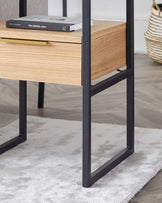 Modern minimalist wooden side table with sleek black metal frame, featuring an open shelf and a drawer for storage. The table is showcased on a soft, light grey rug in a room with light hardwood flooring.