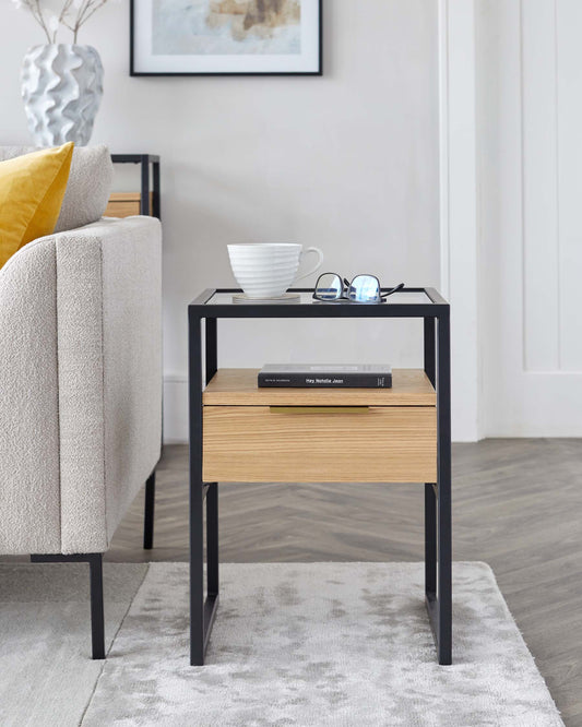 Modern side table with a black metal frame and a light wood drawer, featuring a white ceramic cup, reading glasses, and a hardcover book on its top.