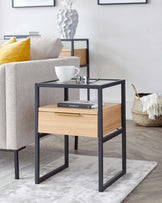 A modern side table with a sleek black metal frame and a light wooden drawer. The table is accessorized with a white cup and saucer, reading glasses, and a book, and it is situated on a soft grey rug. In the background is a textured grey fabric sofa with a bright yellow throw pillow, a decorative vase on a shelf, and a framed artwork on the wall.