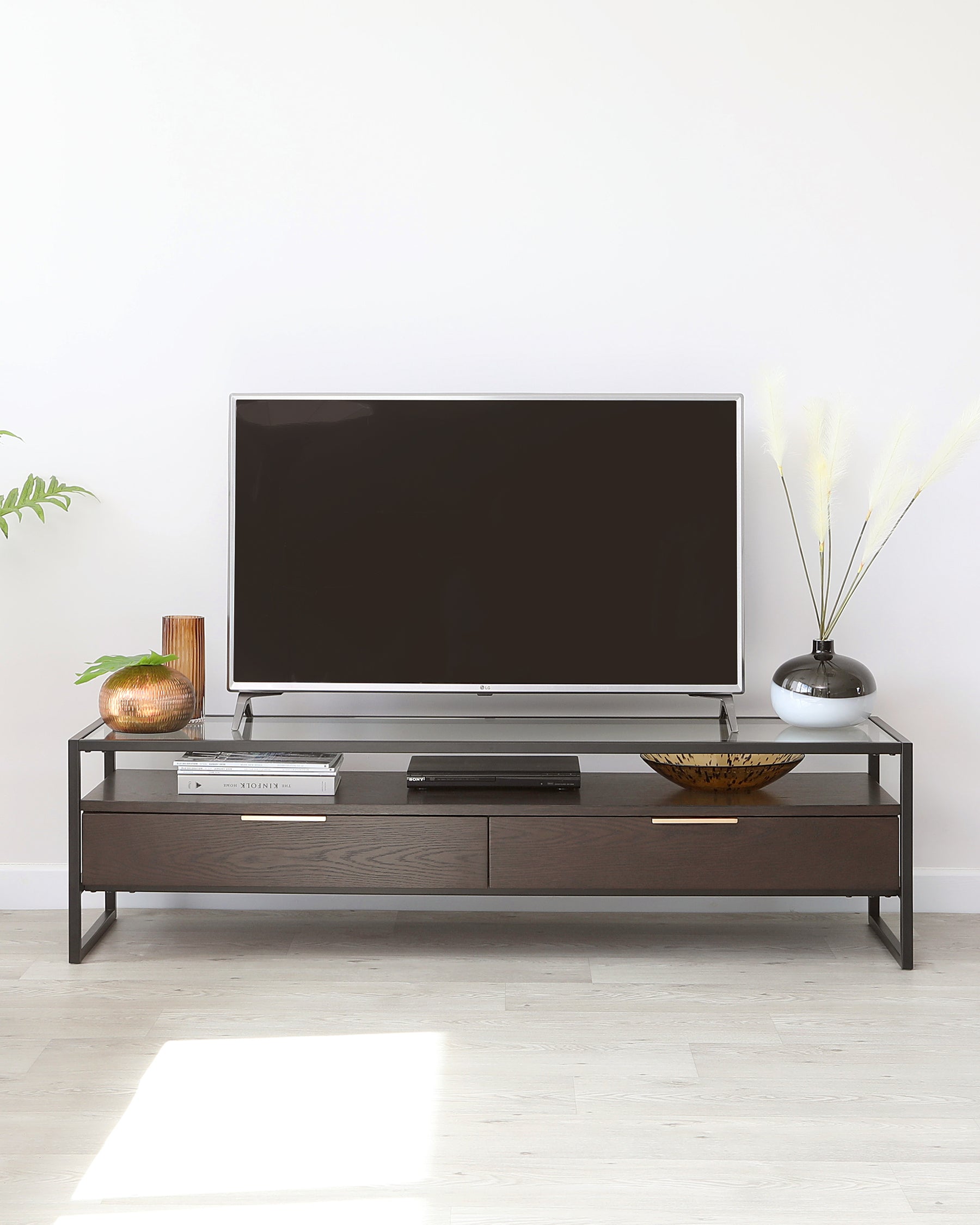 Modern minimalist TV stand with a dark wood finish and metal frame, featuring two drawers and an open shelf for storage, displayed in a bright room with decorative items.