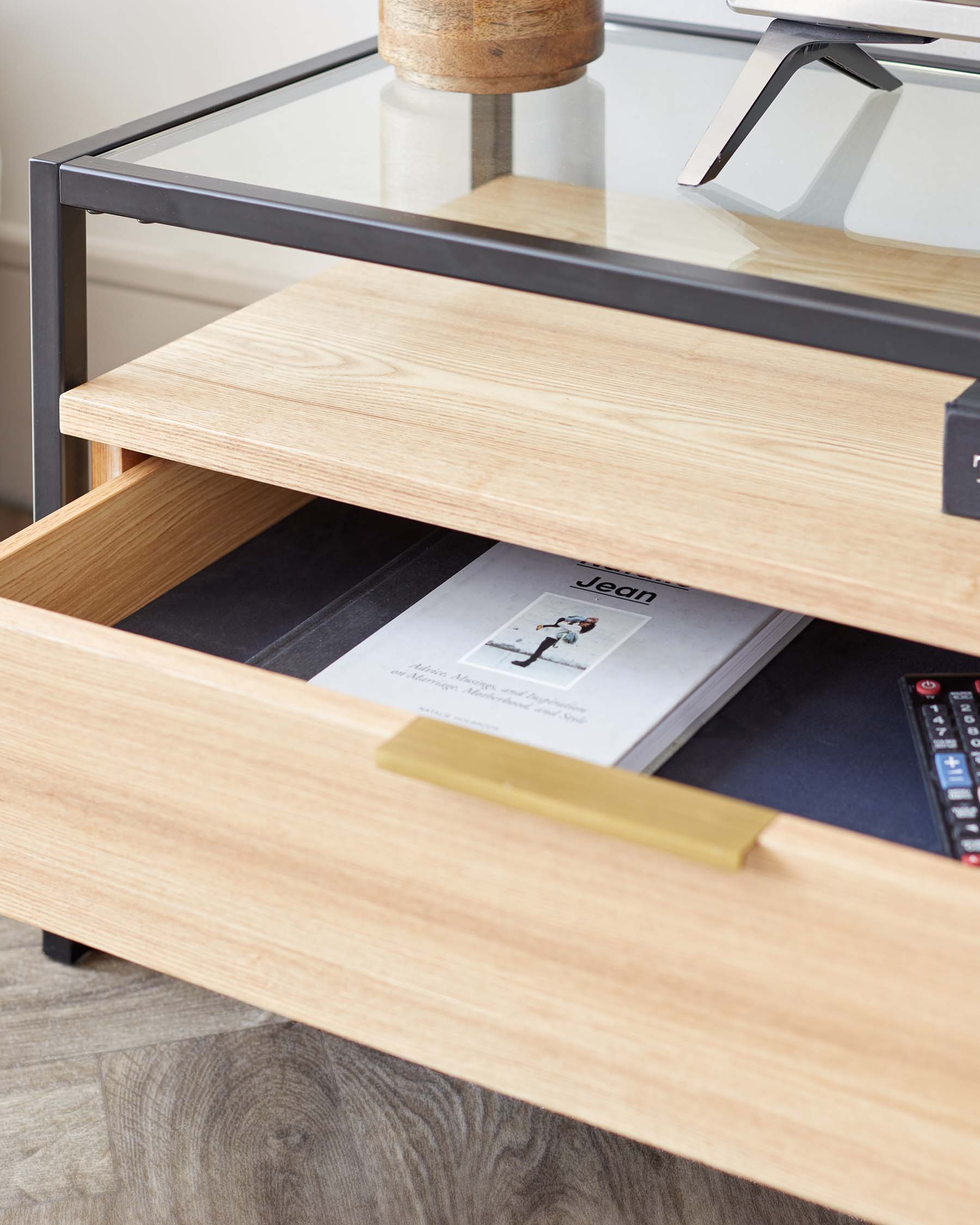 Modern wooden side table with an open drawer revealing a magazine and remote controls, featuring a sleek brass handle, glass top, and black metal frame.