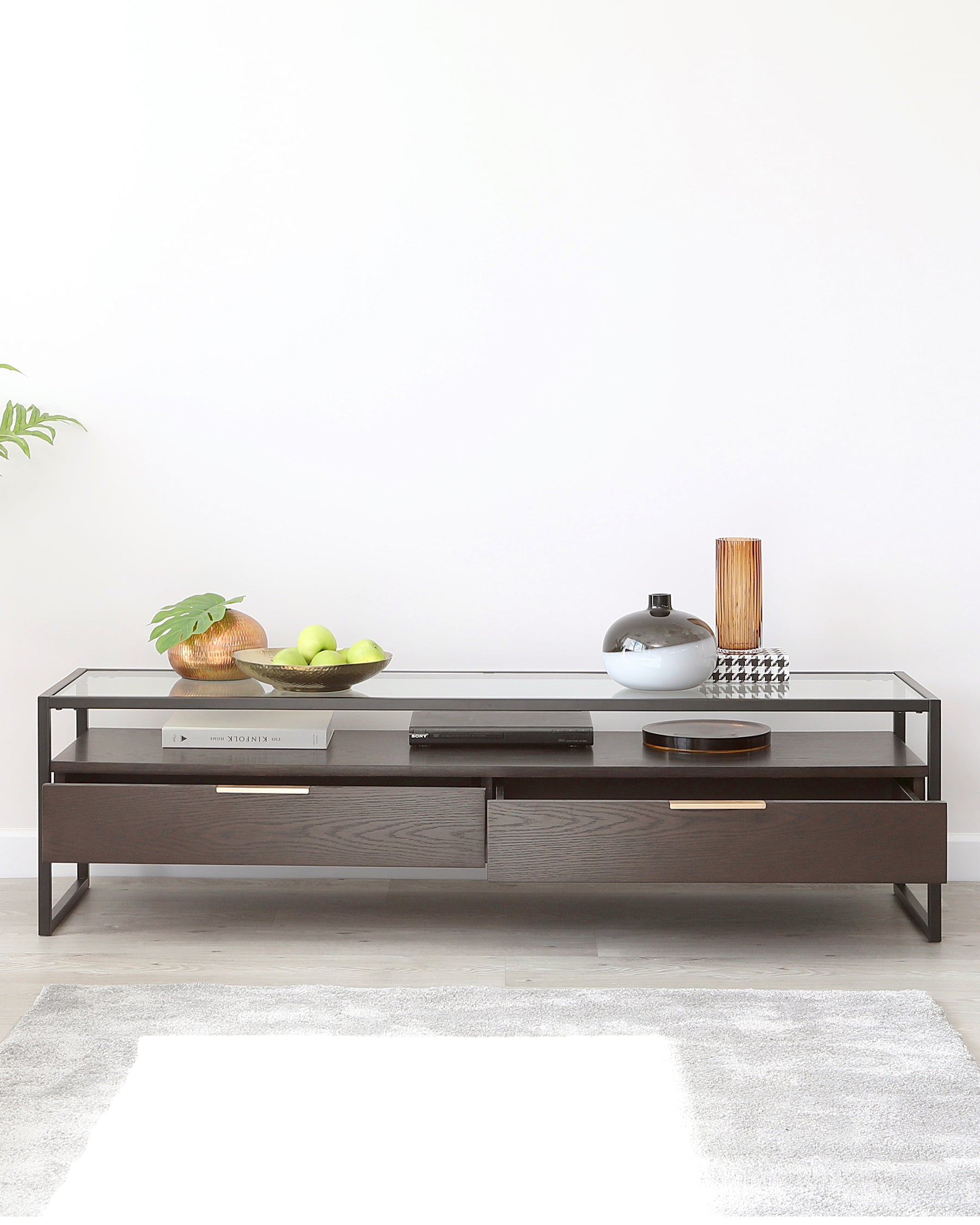 Modern minimalist coffee table featuring a dark wood finish with two pull-out drawers and an open shelf below a glass tabletop, accented with metallic handles and frame, displayed in a bright room with decor items on top and a small area rug beneath.