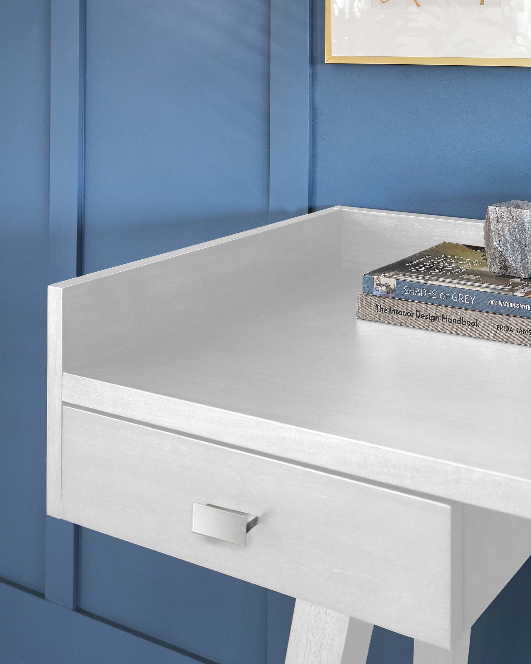 Modern minimalist light grey desk or console table with clean lines, featuring a rectangular tabletop and a single flush drawer with a silver-tone rectangular pull handle. The desk stands on four straight, squared legs, set against a blue panelled wall with a framed artwork partially visible above it. The table is styled with stacked design books on the surface.