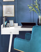 A modern white desk with a sleek design featuring a single centred drawer and angled legs, paired with a luxurious teal blue velvet accent chair that has a high back and subtle gold metal detailing on the base. The elegant pieces are juxtaposed against a deep blue panelled wall, with decorative items and artwork adding a sophisticated touch to the scene.