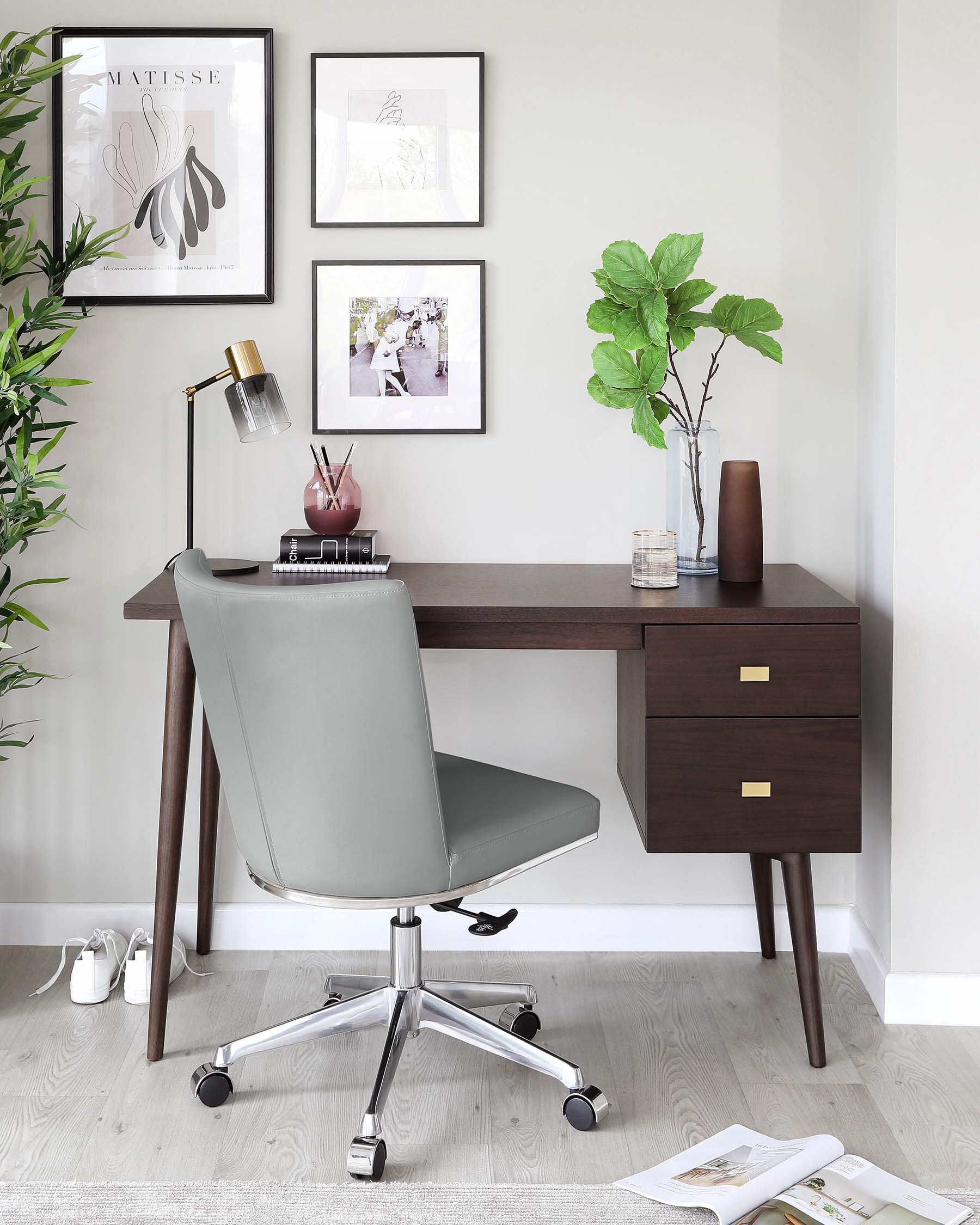 Modern minimalist home office setup featuring a sleek, dark wood finish desk with two drawers featuring brass handles, angled legs matching the desk, and a comfortably padded grey office chair with a minimalist design on a five-point chrome base with casters.