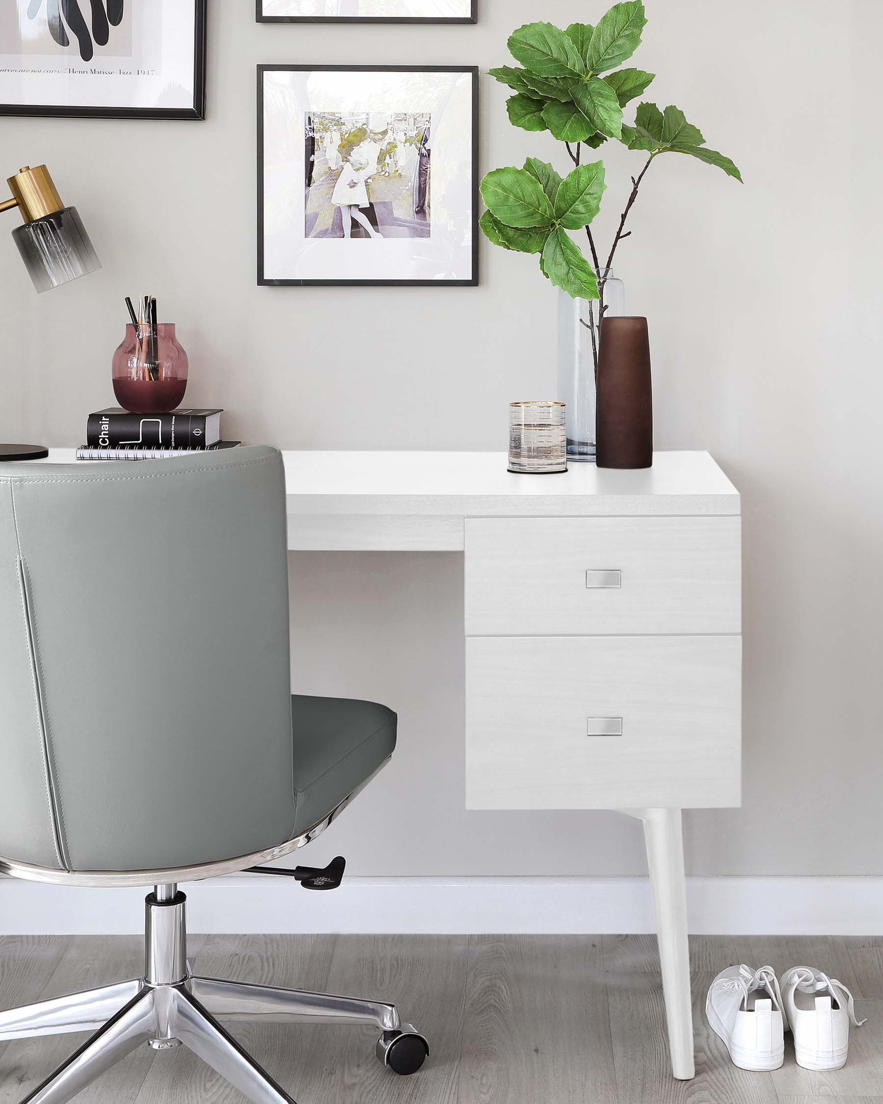 Modern minimalist home office space featuring a white wall-mounted desk with two drawers and a unique supporting leg, paired with a sophisticated grey office chair on a five-point chrome base with casters. The scene is complemented by a potted plant, decorative vases, and framed artwork, creating a clean and inviting workspace.