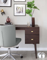 Modern minimalist wooden desk with dark walnut finish featuring two drawers with brass rectangular pull handles, supported by tapered legs. A contemporary chrome-base swivel chair with a grey upholstered seat is placed in front of the desk.