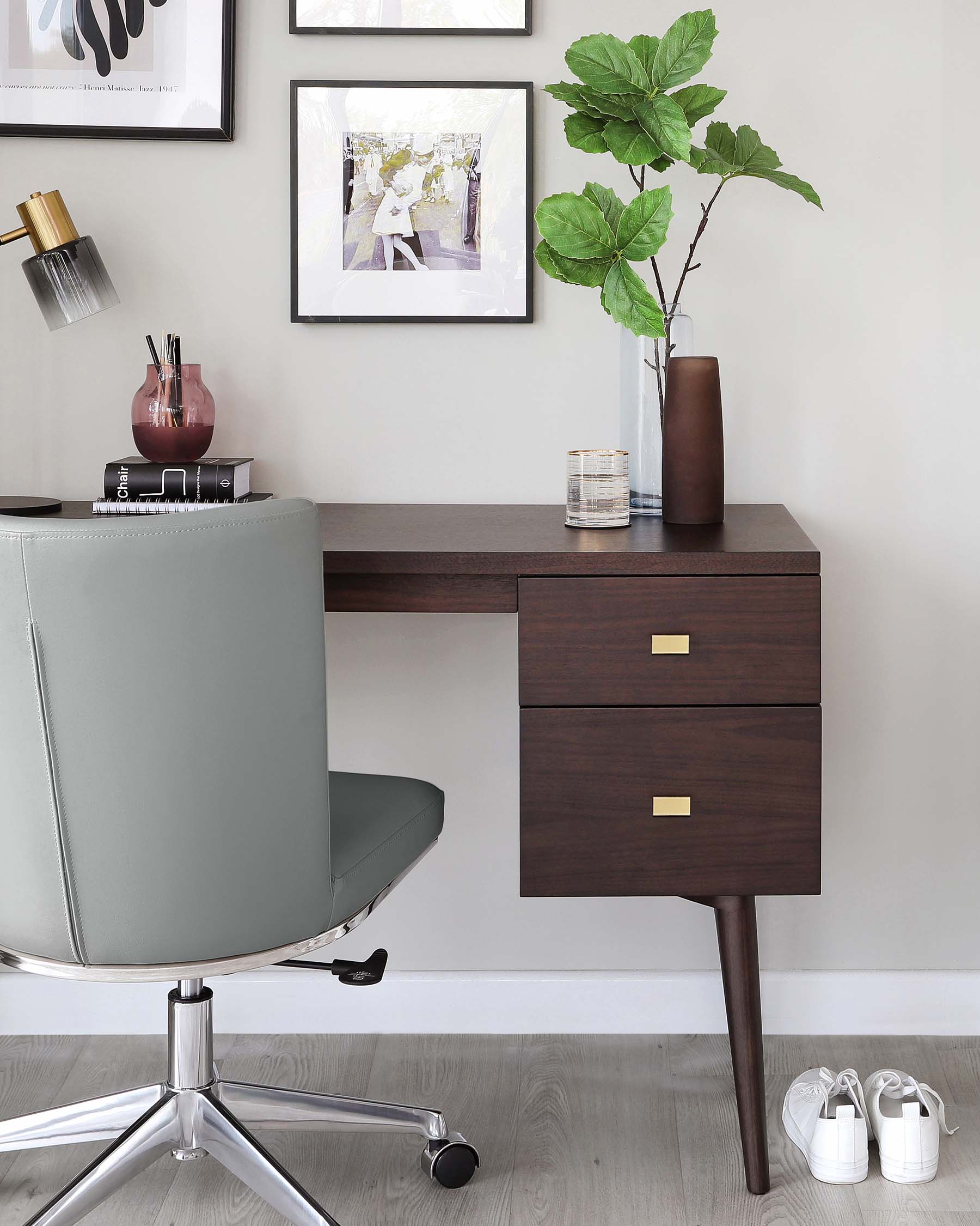 Modern minimalist wooden desk with dark walnut finish featuring two drawers with brass rectangular pull handles, supported by tapered legs. A contemporary chrome-base swivel chair with a grey upholstered seat is placed in front of the desk.