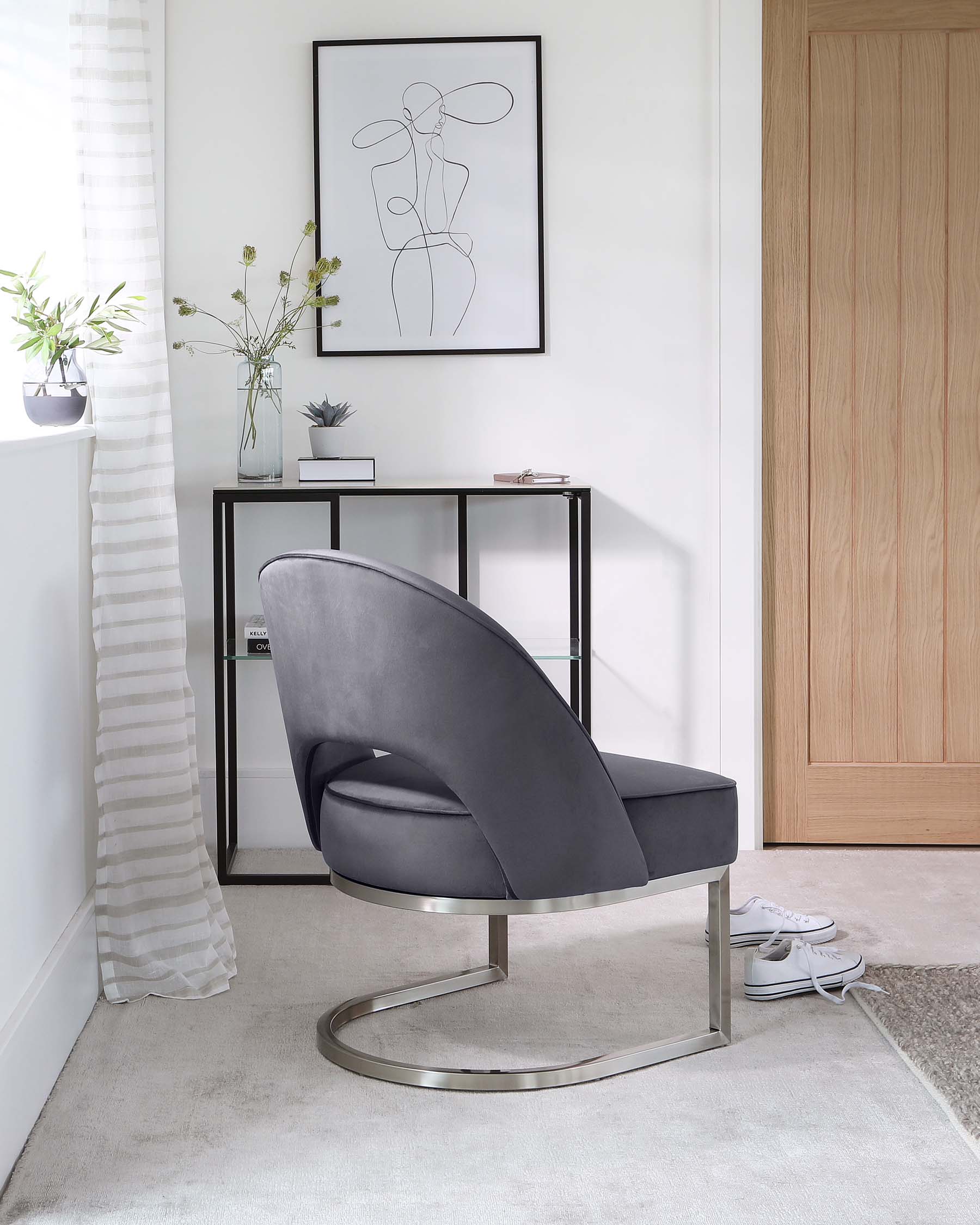 Modern curved lounge chair with dark grey upholstery and a unique silver metal base that forms a continuous curved line for support. In the background, a minimalistic black console table with a glass top, displaying decorative items, complements the chair. Both pieces blend seamlessly with the contemporary room decor.