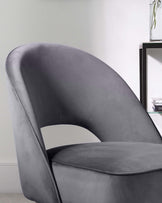 Elegant modern armchair with a smooth curved backrest and thick seat cushion, upholstered in a soft grey velvet fabric. The room setting includes a sleek black-framed side table with glass shelves and a decorative plant on the top shelf.