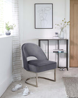 A modern grey upholstered chair with a curved backrest, a cut-out detail on the back, and a unique, symmetrical metal base in a matte finish. Also featured is a sleek, narrow black metal console table with glass shelves, displaying decorative items and books.