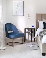 A luxurious velvet blue accent chair with a unique U-shaped metal base, complementing a modern nightstand with a glass top, metal frame, and a lower shelf housing books. A stylish bed with an upholstered headboard is partially visible, adorned with decorative pillows and a throw blanket.