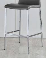 Modern bar stool with a dark grey leather seat and a polished chrome base with slender legs and footrest, on a light wooden floor.