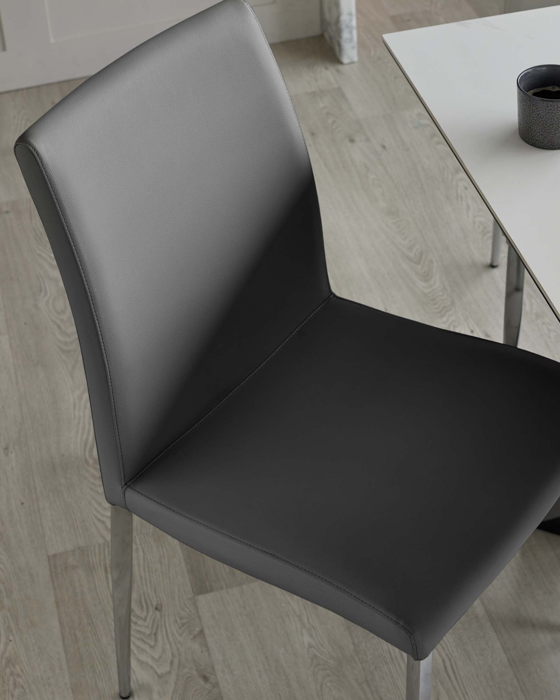 Modern minimalist dining chair made of black faux leather with a high backrest and slender metal legs, paired with a section of a sleek white dining table visible in the top right corner.