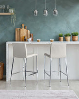 Elegant modern kitchen bar setup featuring two light beige upholstered bar stools with sleek chrome legs, complementing a white marble countertop island with a subtle grey vein pattern.