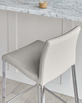 Elegant modern bar stool with a light grey faux leather upholstery and sleek chrome-finished metal legs, positioned beside a white marble countertop with a glass of coffee on top.