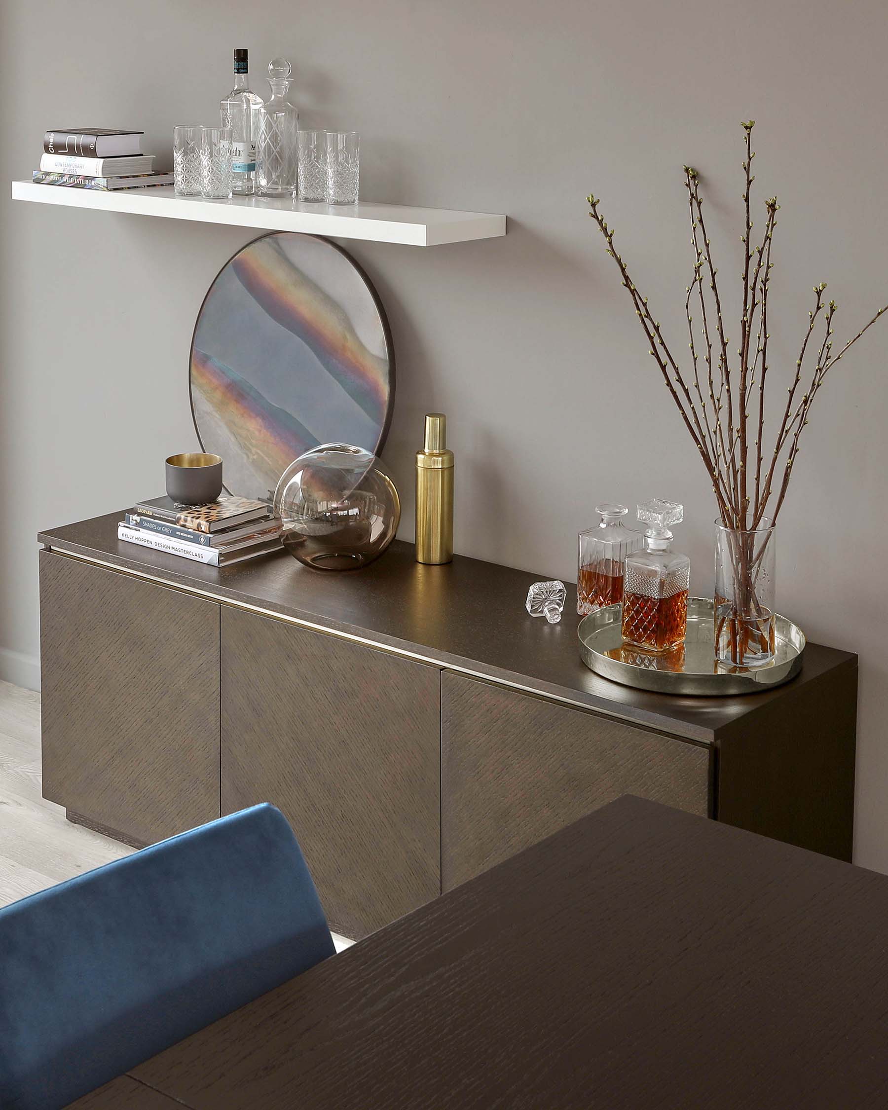 A modern sideboard in a textured grey finish with a metallic countertop, featuring a streamlined design with hidden handles on cabinet doors. A simple white wall-mounted shelf is placed above it.