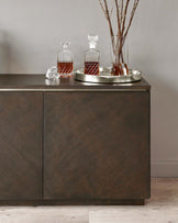 Modern dark wood sideboard with a smooth finish and clean lines, featuring two cabinet doors without visible handles, placed against a light grey wall on a light wood floor. The top displays a silver tray with crystal decanters and whiskey glasses, alongside a tall vase with decorative branches.