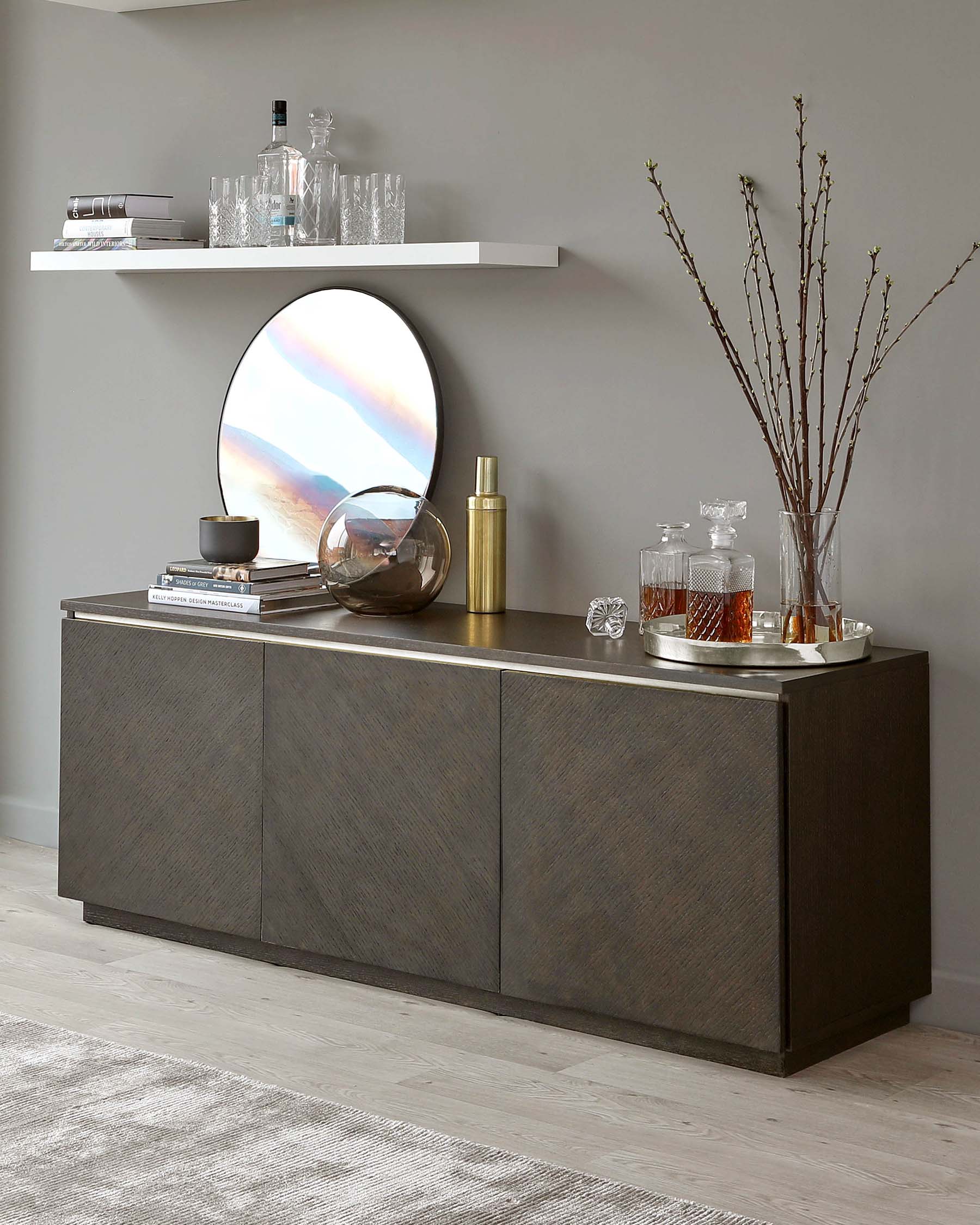 Modern dark wood sideboard with clean lines and handle-less design, featuring a wall-mounted white shelf above and accessorized with a circular mirror, decorative vases, and a selection of decanters on top.