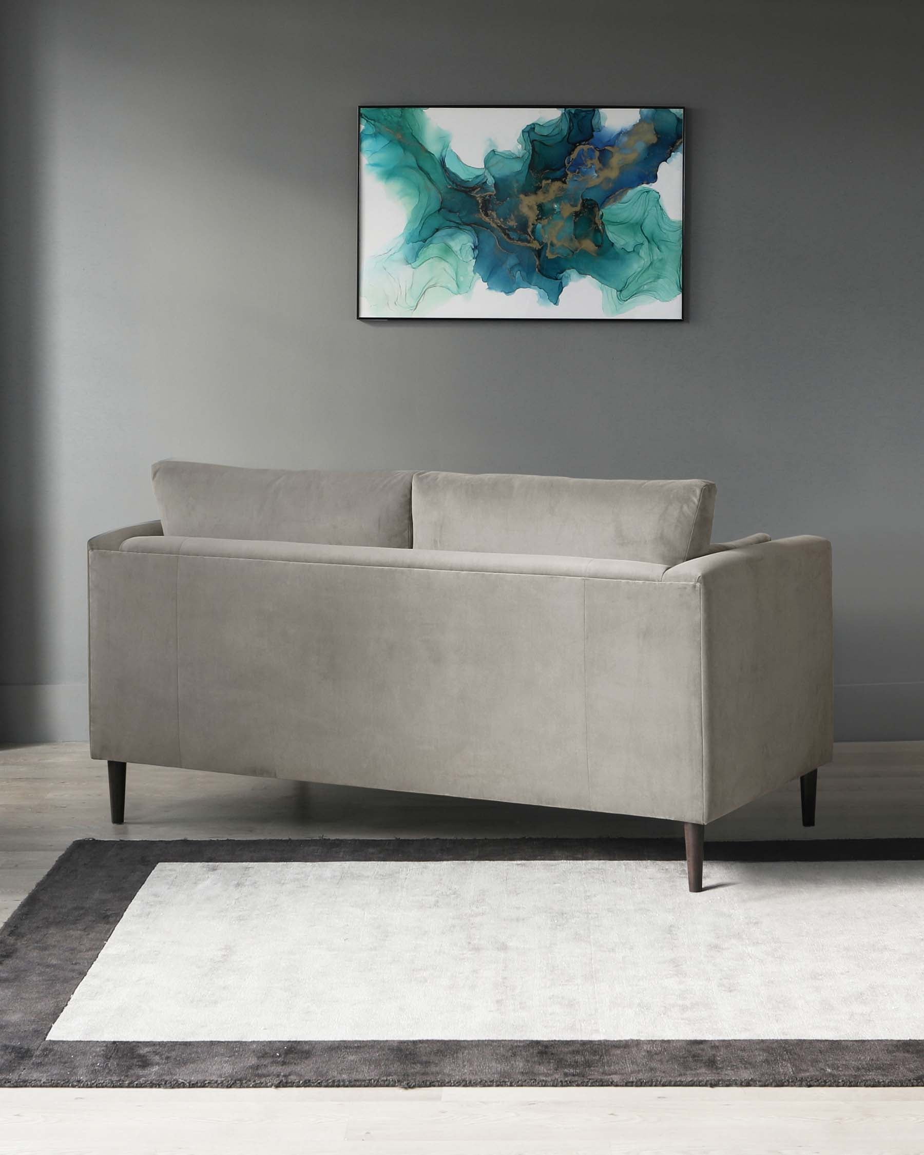 A modern light grey velvet sofa with clean lines and square arms, set on a two-tone grey and white area rug. The sofa is positioned before a grey wall with an abstract blue and green art piece mounted above.