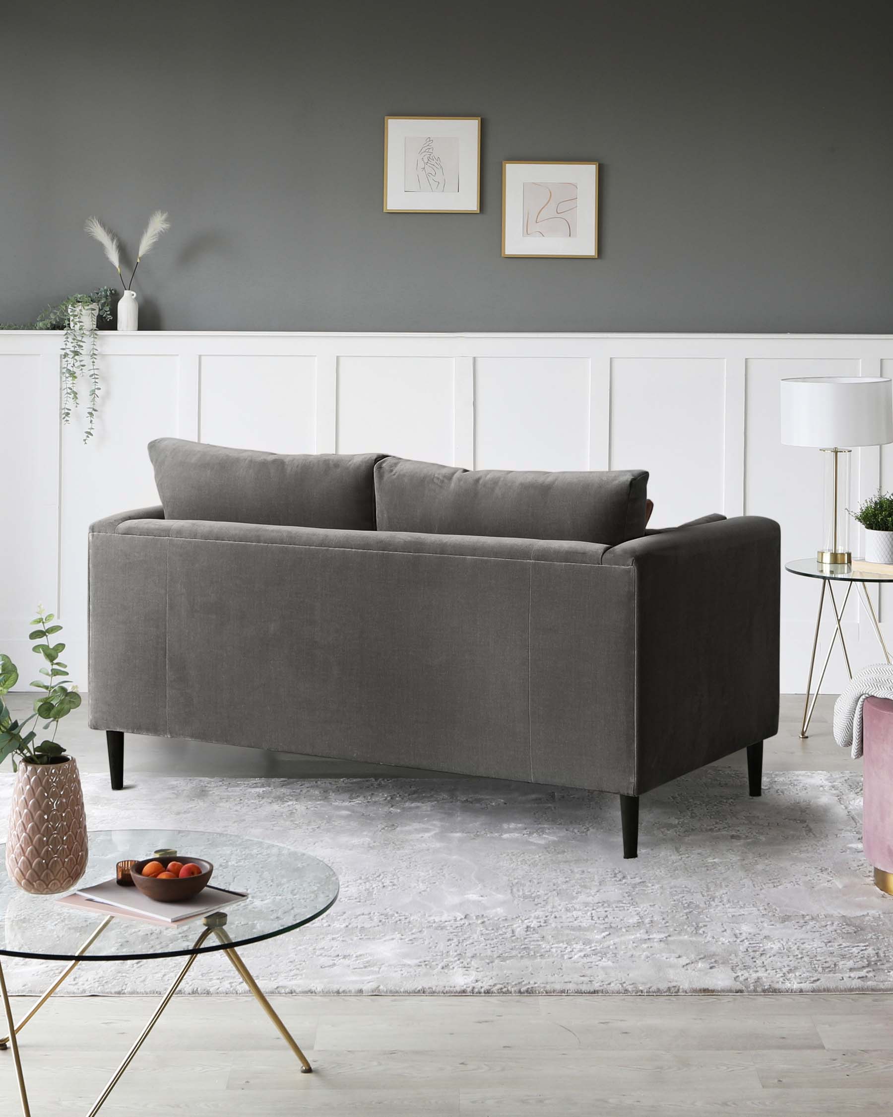 A modern grey fabric two-seater sofa with clean lines and black legs is the centrepiece of the room. In front of it is a round glass coffee table with gold-coloured metal legs. To the side of the sofa, there is a small, round, white marble-topped side table with a gold-coloured metal base, upon which a contemporary white lamp with a gold base and a white lampshade is placed. The furniture is arranged on a white and grey patterned area rug.