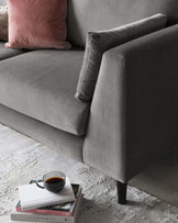 Elegant contemporary sofa in a charcoal grey fabric with a smooth finish, featuring clean lines and minimalist design. A plush pink pillow adds a touch of colour, resting against the structured, cushioned armrest. The couch sits on sleek, dark wooden legs, with a small white side table nearby showcasing a stack of books and a cup of coffee on top. The scene is completed by a textured grey and white area rug underneath.