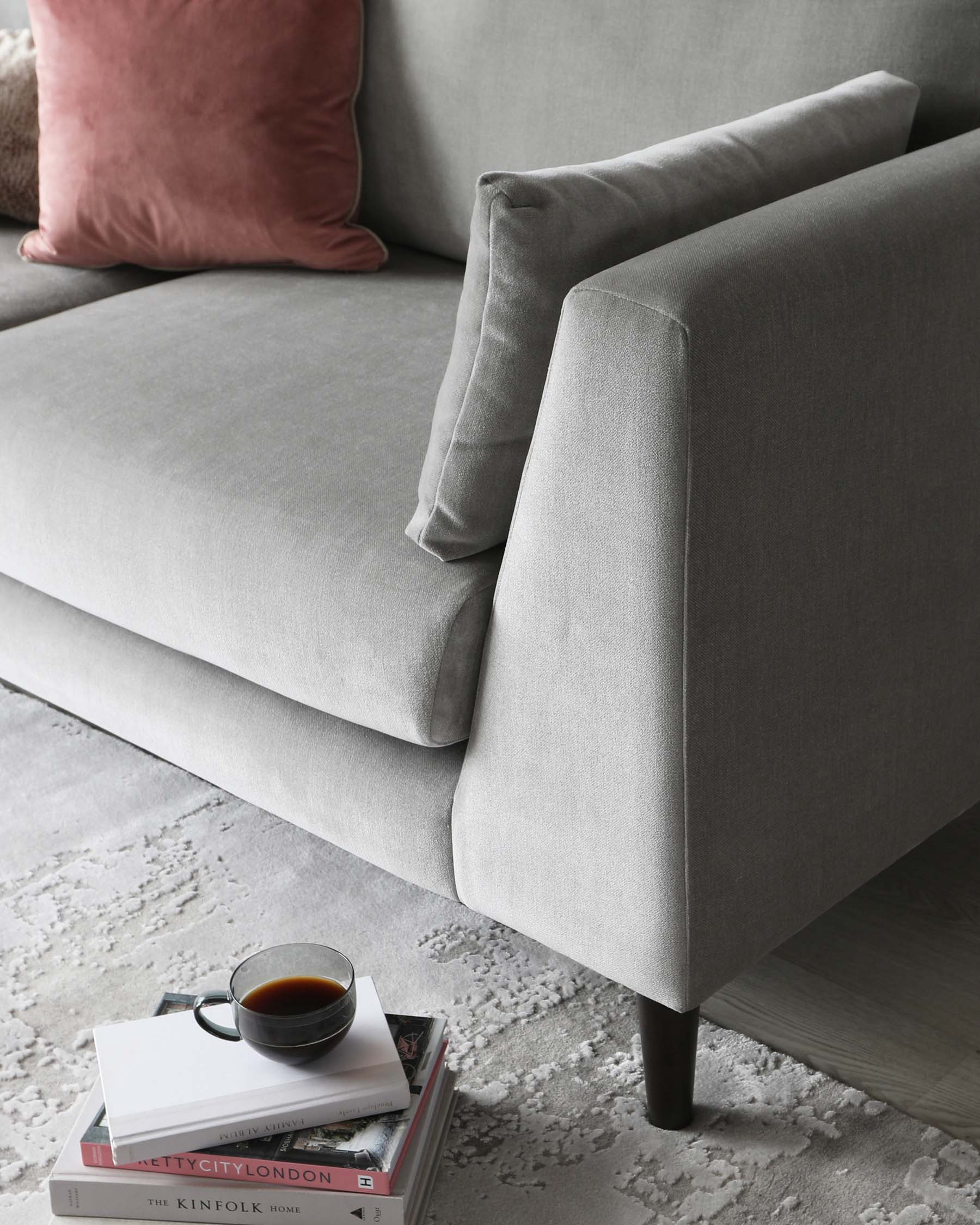 A contemporary grey fabric sofa with clean lines and a minimalist design, featuring a square armrest and plush cushioning. Accented with a rose-coloured pillow, the sofa is complemented by a stack of books and a cup of coffee on the floor, indicating a cosy, lived-in atmosphere.