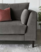 A contemporary charcoal grey fabric sofa with plush cushions, featuring a solid backrest, one cylindrical side cushion, and a rectangular pink accent pillow. The couch stands on dark wooden legs and is complemented by a soft, textured rug underneath.