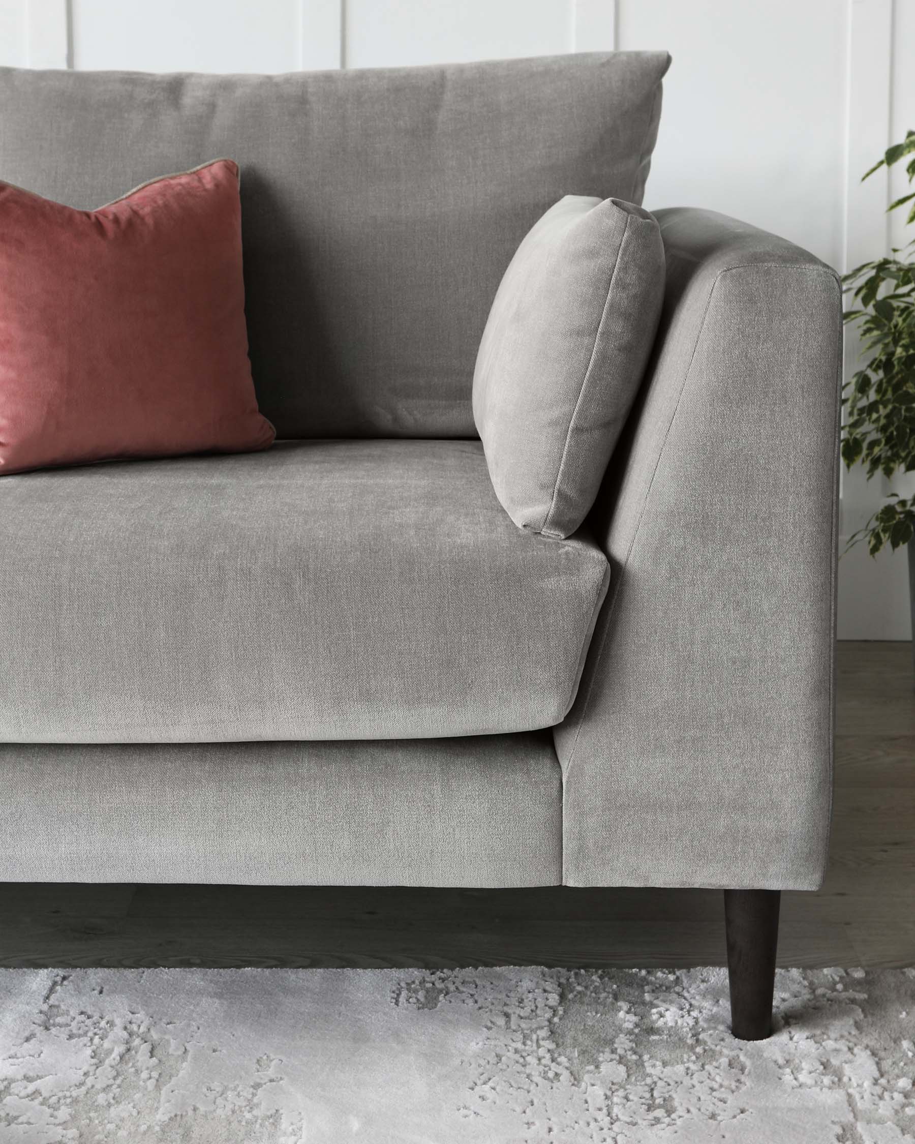 Gray fabric sofa with a streamlined modern design, featuring clean lines and dark wooden legs, complemented by a soft pink accent cushion. Visible in the backdrop is a plush white rug with a subtle pattern, set against a light wooden floor and white wall, which adds to the contemporary and cosy aesthetic of the scene.